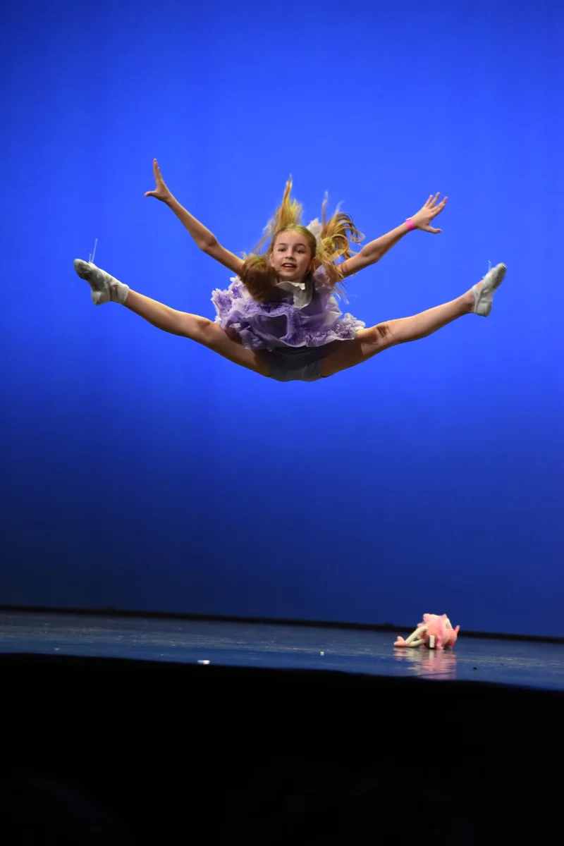 Una ballerina esegue una spaccata in salto davanti a uno sfondo blu.