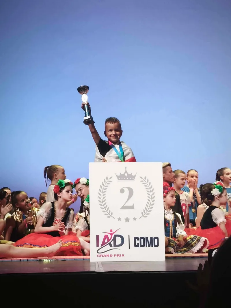 Un bambino solleva un trofeo sul podio circondato da giovani ballerini.